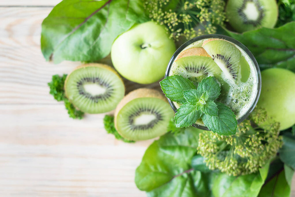 Green smoothie in a glass, decorated with mint and kiwi, with apples, kiwi, cucumbers and parsley in background, selective focus, top view - 写真・画像