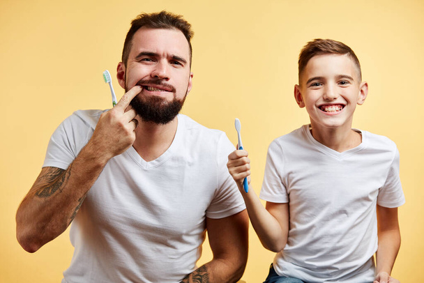 father and son brushing teeth and looking at camera together isolated on yellow - Foto, Imagen