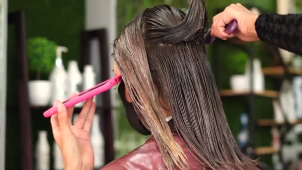 A girl hairdresser makes a client keratin hair straightening. The girl is sitting in a black medical mask in the salon. The master applies the straightening product to the hair    - Footage, Video