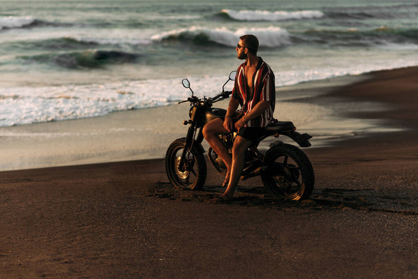 ビーチで自転車を持って魅力的な孤独な男が夕日を満たしています。海のそばで自転車でスタイリッシュな男。自転車で旅行しなさい。バリへの旅行。熱帯のビーチでのカスタムバイク. - 写真・画像