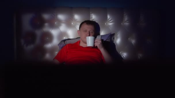 Een jonge man ligt 's nachts op het bed met een mok in zijn handen en kijkt tv, camera beweging - Video