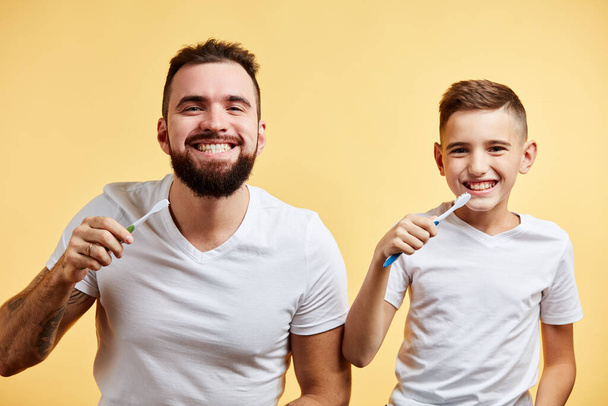 father and son brushing teeth and looking at camera together isolated on yellow - Fotó, kép