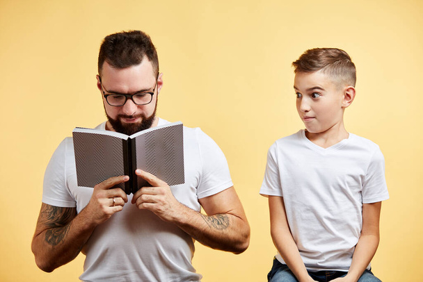 father incredulously looking at cute little son reading book at yellow background - Φωτογραφία, εικόνα