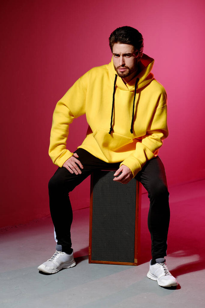 A young man of 25-30 years old in a yellow sweatshirt sitting on an old music column on pink wall background and showing different emotion. - Фото, изображение
