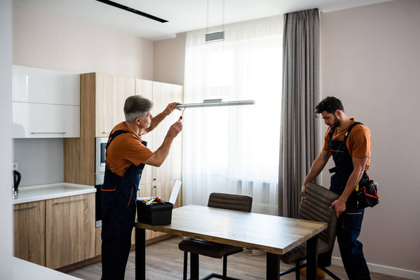 We make it better. Two handymen, workers in uniform fixing, installing light lamp and equipment in the kitchen, using screwdriver indoors. Repair and assembly service concept - Zdjęcie, obraz