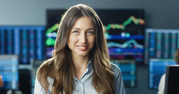 Close up portrait of happy female trader or broker working at stock exchange office looking at camera on background of multiple monitors showing data, ticker numbers and graphs. - Photo, Image