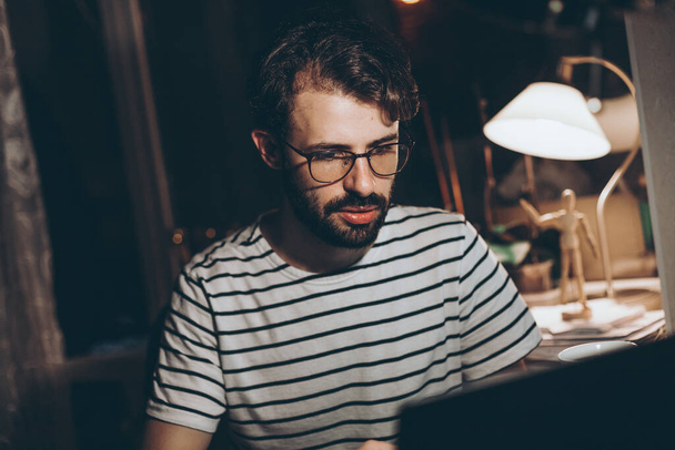 portrait d'un jeune homme travaillant à la maison en ligne à l'ordinateur le soir - Photo, image