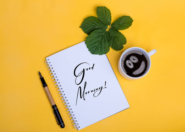 A cup of coffee with smile, notebook with the sign,  ink pen and leaf on yellow background - Photo, Image