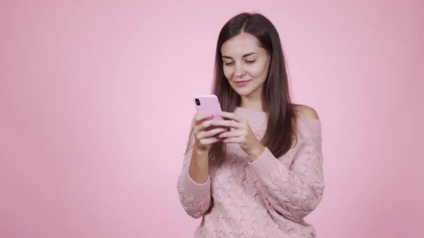 Portrait of woman in pink sweater uses mobile phone, isolated on pink background - Filmagem, Vídeo