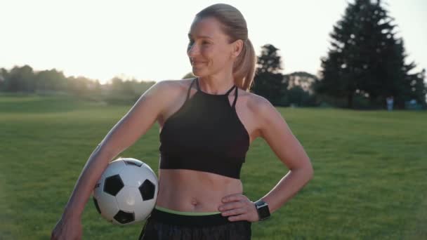 Ready to play. Portrait of attractive sporty woman holding soccer ball in hand, looking at camera and smiling while standing in green field on a summer morning - Footage, Video