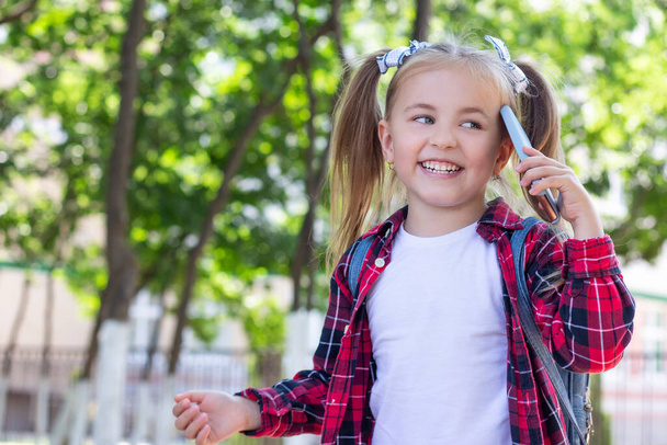 happy schoolgirl with a backpack talking on the phone on the street. in a white T-shirt and a plaid shirt - Foto, Imagem