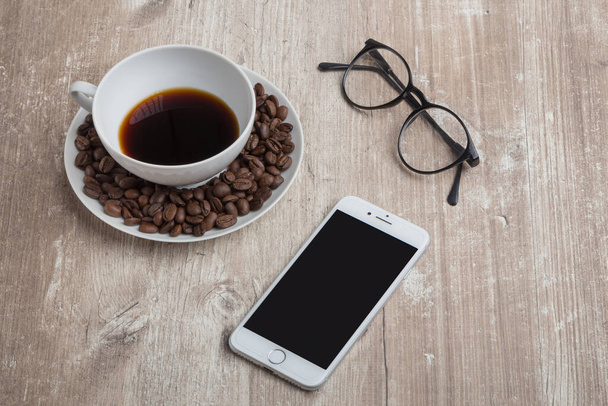 a cup with coffee beans, a white phone and reading glasses on an old textured wooden table - Photo, Image