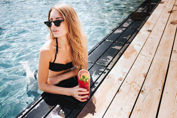 Jovem mulher quente descansando na piscina. Menina em biquíni escuro sexy e óculos de sol segurando coquetel vermelho e posando na câmera. Descansar no resort hotel spa lugar. Em pé na água
. - Foto, Imagem
