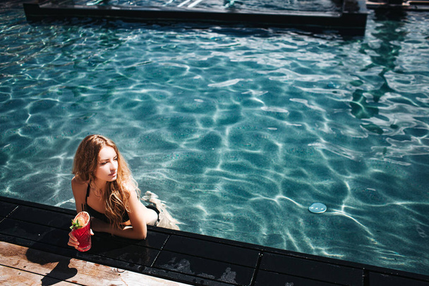 Молодая горячая женщина отдыхает в купальнике. Фото девушки в воде, позирующей в одиночестве. Женщина держит красный коктейль в руке и смотрит в сторону. Сексуальная девушка в курортном отеле. - Фото, изображение