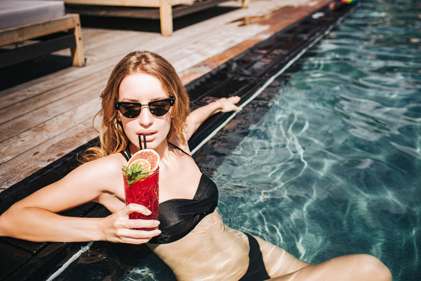 Young hot woman resting at swimpool. Picture of well-built slim and sexy girl drinking red cocktail and enjoy her rest in clear water. Hotel resort spa. Summer sunshine. - Foto, Imagen