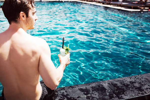 Young hot man resting at swimpool. Cut back view of strong powerful guy holding cocktail in hand and enjoy spending time at water. Summertime vacation. - 写真・画像