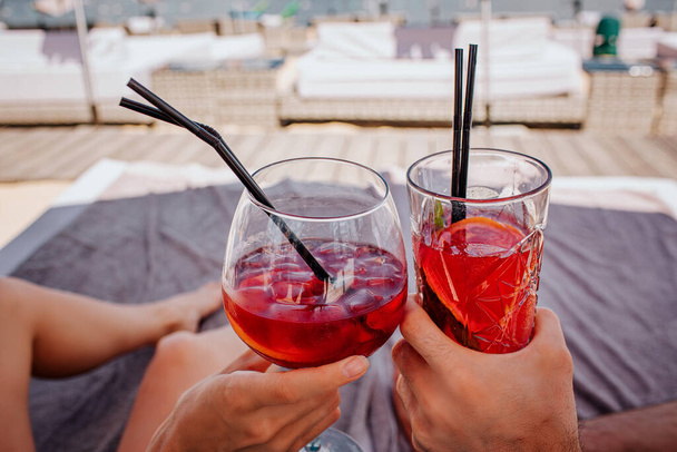 Junges heißes Paar, das sich am Swimmingpool ausruht. Bild von zwei roten kalten Cocktails mit Eiswürfeln, die jubeln. Entspannung und Erholung während des Urlaubs in den Sommertagen. Kurzeit. - Foto, Bild