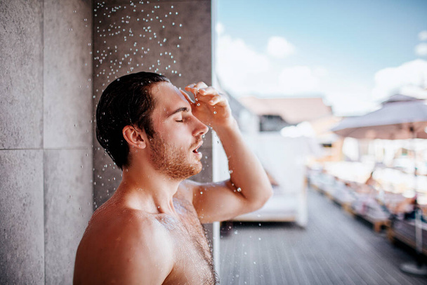 外でシャワーを浴びてる若い男。シャワーの下にシャツなしで男のサイドビューの肖像画ながら、水が落ちてくる。水の手順を楽しむ. - 写真・画像