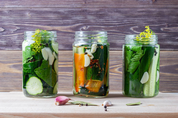 Homemade pickled vegetables in a jars: cucumbers, paprica and garlic with herbs on a wooden background. The canned or fermented vegetarian food concept. - Φωτογραφία, εικόνα