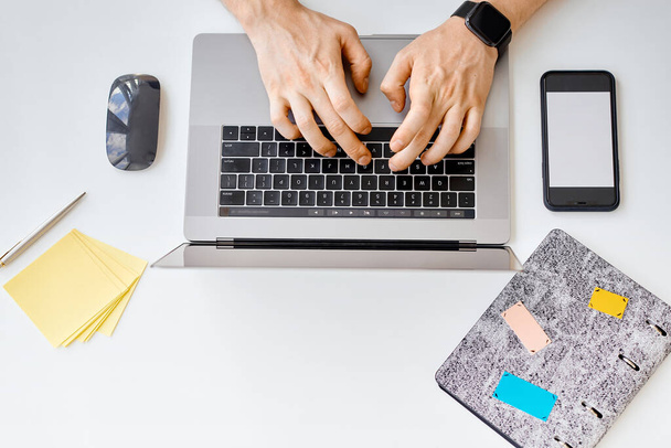 Een man typt tekst, werkt achter een laptop, naast een telefoon met een witte kraan  - Foto, afbeelding