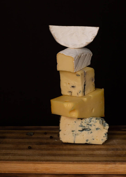 Koyu arkaplandaki ahşap kesme tahtasındaki farklı peynir türleri. Dikey. Boşluğu kopyala - Fotoğraf, Görsel