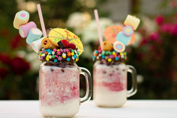 Freakshakes in jars with cocktail umbrella, straw, marshmallow, strawberry, chocolate on a white table on a background of summer flowers. - Photo, Image