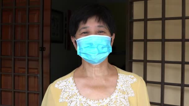 Elderly Chinese woman, face covered with medical face mask. Standing outside her house. - Filmati, video