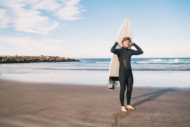 Portrait de jeune surfeur à la plage tenant sa planche de surf et portant un costume de surf noir. Sport et sport nautique concept. - Photo, image