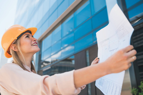 Portrait of professional architect wearing yellow helmet and looking at blue prints outside modern building. Engineer and architect concept. - Photo, Image