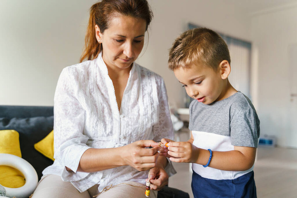 Front view on small caucasian boy 4 years old making bracelet with his mother - Little child playing with his mother learning new skills creative development at home in day - trust and support concept - Photo, Image