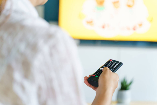 Back view on hand of unknown caucasian woman holding remote controller changing TV channel at home - Television or internet smart tv choosing content using modern technology leisure relaxation concept - Photo, Image