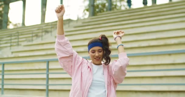 Portret pięknej, radosnej, młodej, białej kobiety w różowej kurtce tańczącej i skaczącej na świeżym powietrzu. Bardzo szczęśliwa dziewczyna w dobrym nastroju, poruszająca rękami w górze i uśmiechnięta na ulicy. Koncepcja retro - Materiał filmowy, wideo