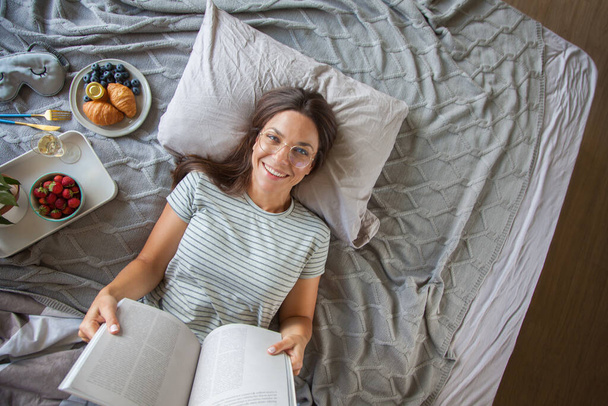 Woman reading book or newspaper and drinking coffee breakfast on bed during the morning - Фото, изображение