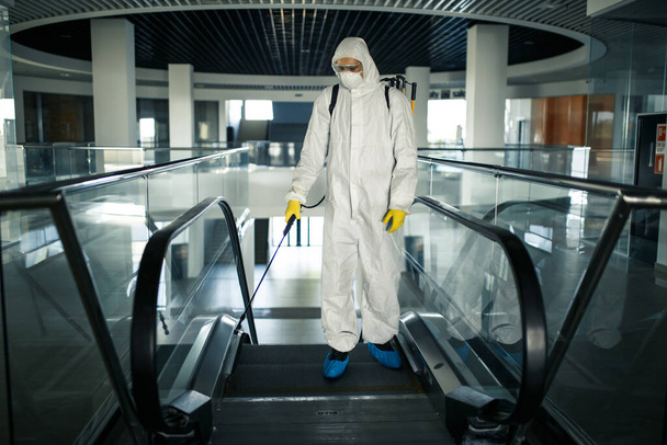 防護服を着た消毒作業員は、専門的に空のビジネスセンターでエスカレーターをクリーンアップします。抗菌服を着た男がショッピングモールを滅菌します。covidの概念 - 写真・画像