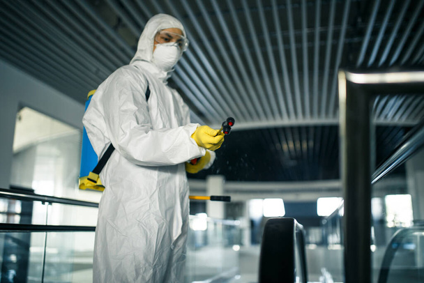 Disinfection worker wearing a protective suit professionally cleans up an escalator in an empty business center. A man equiped with antibacterial clothes sterilizes shopping mall. Covid concept - 写真・画像