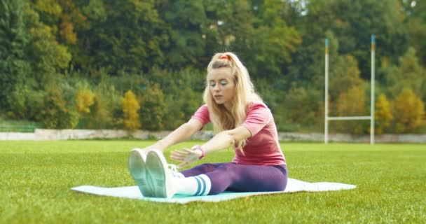Mooie vintage blonde atleet in roze bodysuit opwarmen en doen oefeningen tijdens het zitten. Mooie blanke jonge retro-stijl sportvrouw stretching spieren op yoga mat buiten. Fitness concept - Video