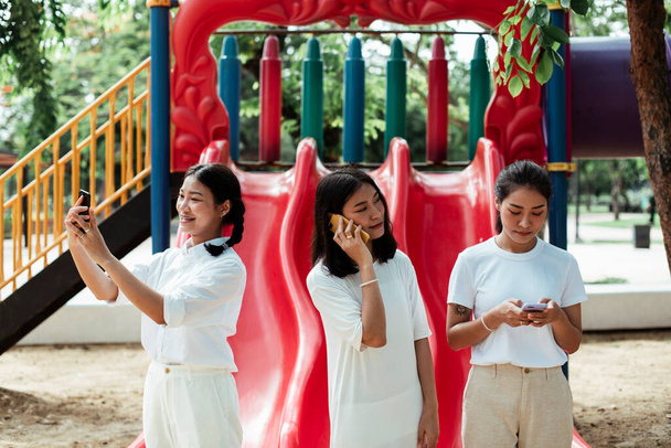 Triple δίδυμη αδελφή στέκεται στο τέλος του κόκκινου slider στην παιδική χαρά, χρησιμοποιούν το δικό τους smartphone. - Φωτογραφία, εικόνα
