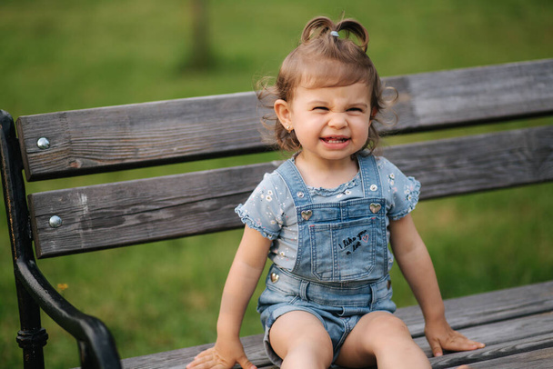 Adorable niñita riéndose. Hermosa niña en denim sentada en el banco en el parque. Feliz sonrisa de niña. dieciocho meses
 - Foto, Imagen