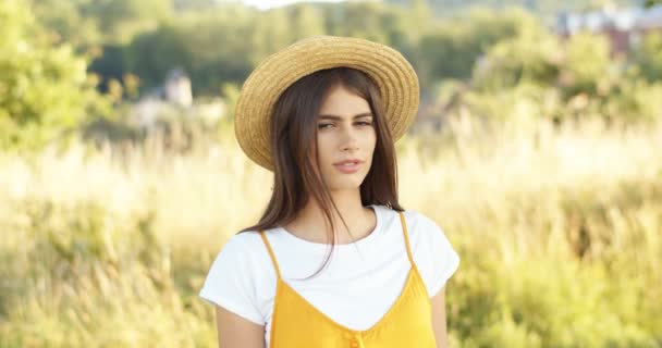 Ritratto di bella giovane bruna donna caucasica in cappello tanding in campo e guardando la fotocamera. Ragazza affascinante con i capelli scuri a posto natura in estate. All'aperto. - Filmati, video