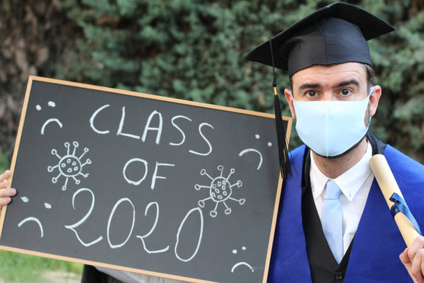 крупным планом портрет красивого молодого студента в выпускной одежде с дипломом и доской класса 2020 в парке - Фото, изображение