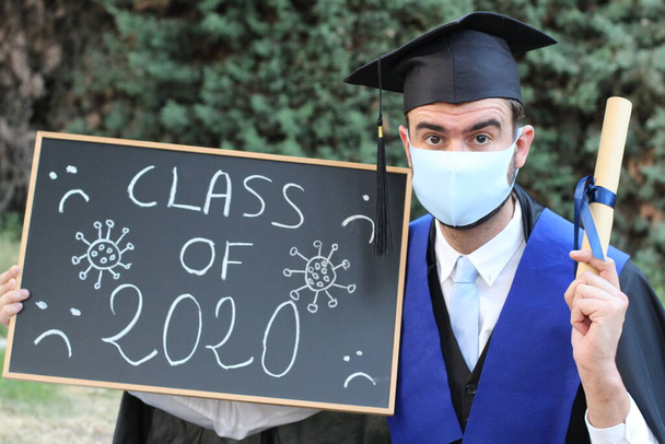 close-up portrait of handsome young student in graduation clothes with rolled diploma and blackboard with class of 2020 inscription in park - Photo, Image