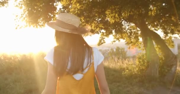 Die Kamera folgt dem schönen und fröhlichen kaukasischen Mädchen mit langen brünetten Haaren und Hut, das bei hellem Sonnenuntergang in einem Feld am Baum spaziert. Hinten. Ziemlich glückliche Frau lächelt in die Kamera und schlendert durch die Natur - Filmmaterial, Video