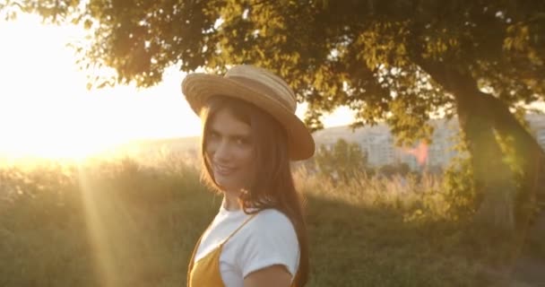 Camera following beautiful joyful Caucasian girl with long dark hair in hat walking in field at tree at bright sunset in suburb. Rear. Pretty happy female smiling and strolling at nature of outskirt. - Footage, Video