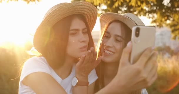 Dos chicas jóvenes y alegres caucásicas en sombreros tomando fotos selfie con cámara de teléfono inteligente a la luz del sol. Feliz hermosa mejor amigos sonriendo al teléfono al hacer la foto en la naturaleza. Fotografía exterior. - Metraje, vídeo