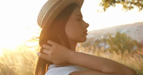Detailní záběr mladé kavkazské krásné dívky v klobouku s dlouhými brunetovými vlasy otáčení tváří v tvář kameře a veselý úsměv při západu slunce na přírodě. Portrét půvabné feny v terénu v létě. - Záběry, video