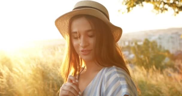 Portrait of young Caucasian beautiful girl in hat with long brunette hair looking at camera like flirting and turning face on sunset at nature. Close up of pretty charming female in field in summer. - Footage, Video