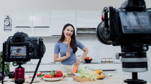 Έννοιες των μέσων κοινωνικής δικτύωσης Μια ευτυχισμένη γυναίκα που στέκεται στην κουζίνα χρησιμοποιώντας μια κάμερα και καταγράφοντας βίντεο online Ευτυχισμένη Ασιάτισσα vlogger μεταδίδει ζωντανά online βίντεο Διδασκαλία μαγειρικής στην κουζίνα στο σπίτι. - Φωτογραφία, εικόνα