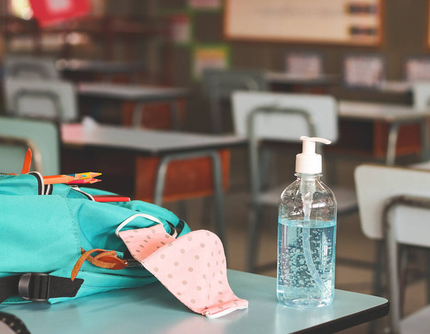 COVID-19 preventie, terug naar school en nieuw normaal concept.Vooraanzicht van ontsmettingsmiddel handgel, roze stippen stof masker en rugzak met schoolbenodigdheden op schoolbureau in de klas. - Foto, afbeelding