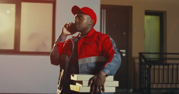 Pizza repartidor afroamericano en el uniforme rojo y la tapa de pie en la casa con cajas de pizza y llamando por teléfono en la noche. Exterior. - Imágenes, Vídeo