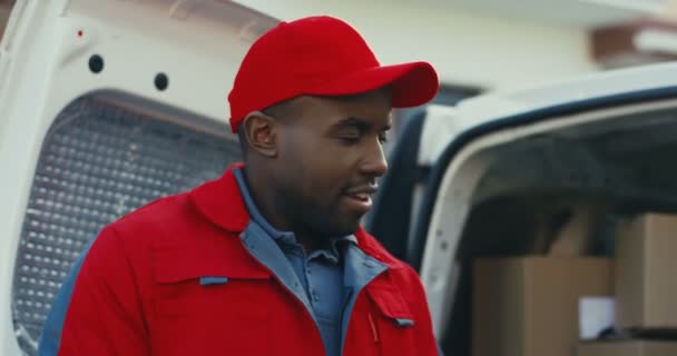 Primer plano del joven repartidor afroamericano con el uniforme rojo y la gorra al aire libre en la furgoneta blanca y contando los buzones. - Imágenes, Vídeo